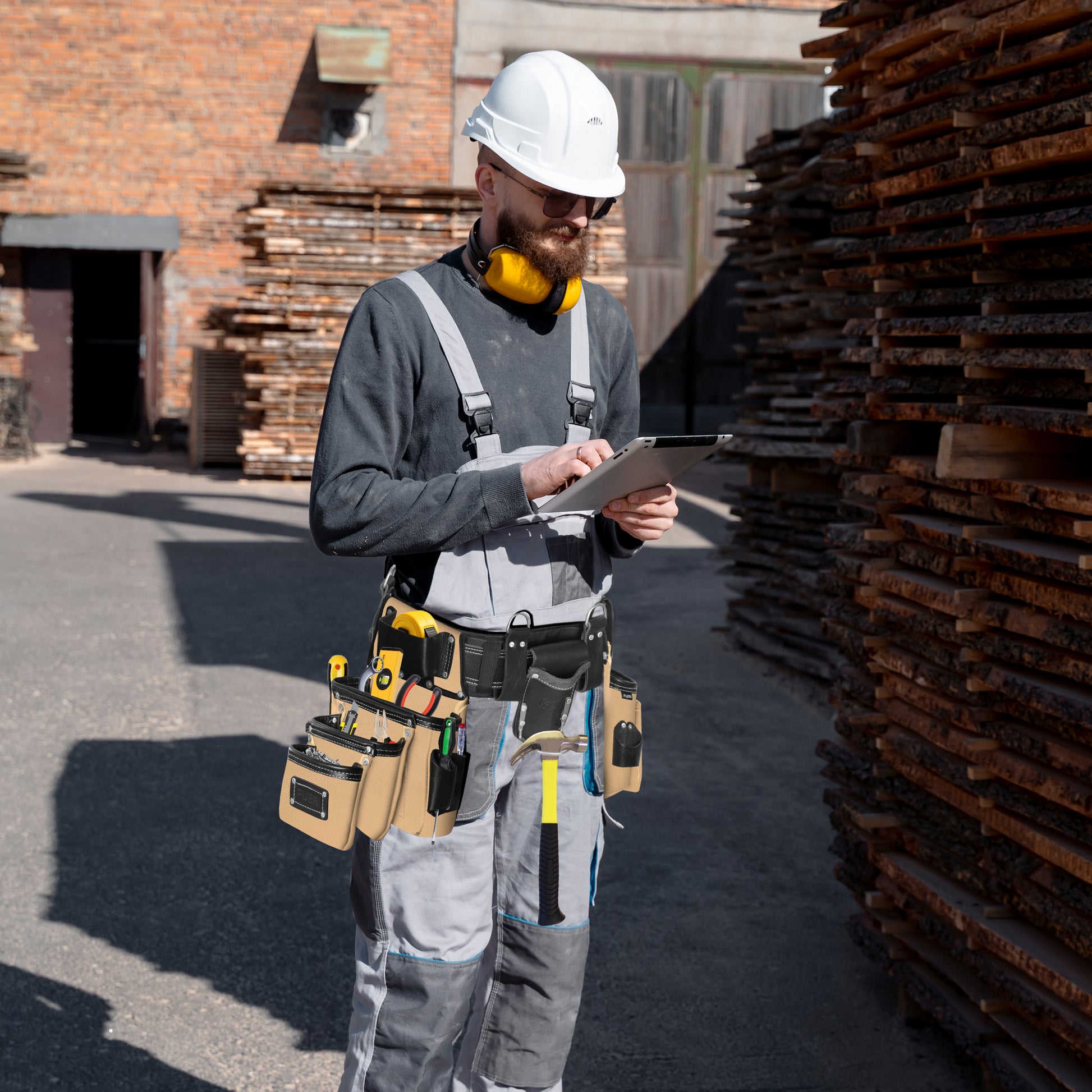 Carpenter in white hard hat and grey overalls wearing a 26-pocket black and beige leather tool belt on a lumber yard jobsite while using a tablet