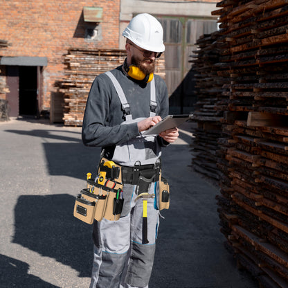 Carpenter in white hard hat and grey overalls wearing a 26-pocket black and beige leather tool belt on a lumber yard jobsite while using a tablet